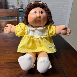 Op de afbeelding: Een vintage Cabbage Patch Kid pop met bruin garen haar, roze wangen en een gele jurk met witte stippen. De pop draagt witte sokken en heeft haar armen uitgestrekt. Een witte strik siert het haar van de pop.