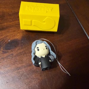May include: A yellow Hallmark "Mystery Ornament" box with a question mark and a small figurine with blue hair and a black outfit. The figurine has a string attached for hanging.