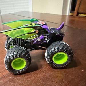 May include: A toy monster truck with a black body, purple accents, and green highlights. The truck has large black tyres with green rims and a green wing-like structure on top. The toy is designed with a dragon-like appearance.