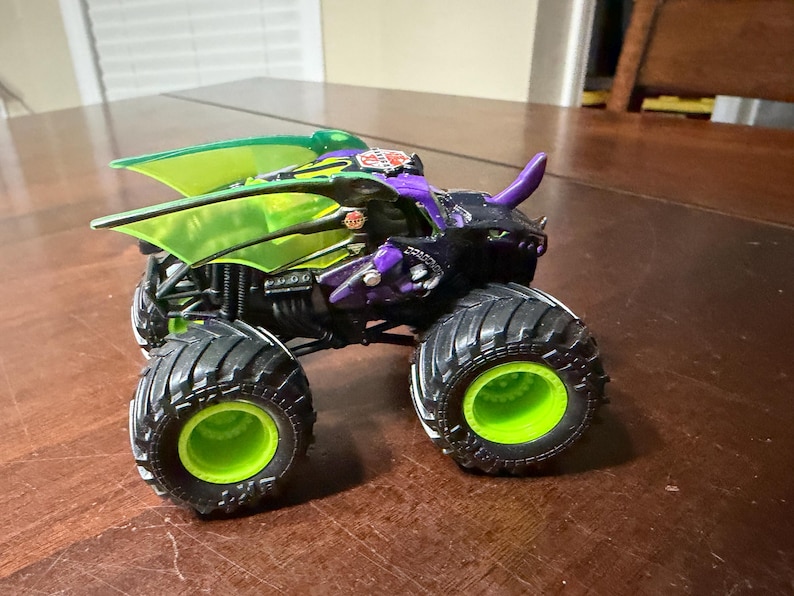May include: A toy monster truck with a black body, purple accents, and green highlights. The truck has large black tyres with green rims and a green wing-like structure on top. The toy is designed with a dragon-like appearance.