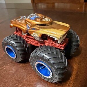 May include: A toy monster truck with a gold and brown body, red chassis, and large black tyres with blue accents. The truck features a detailed design with a menacing front grille.