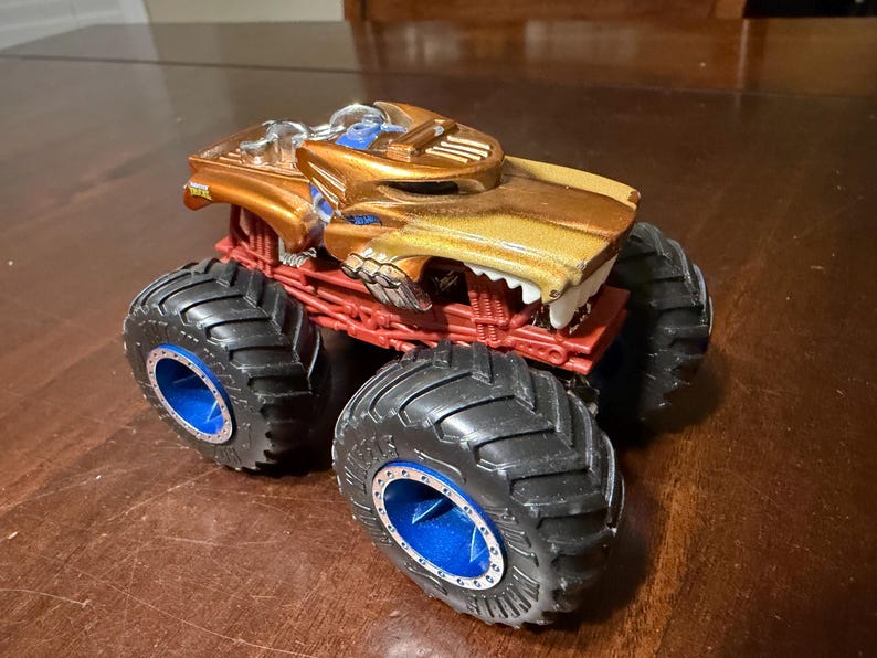 May include: A toy monster truck with a gold and brown body, red chassis, and large black tyres with blue accents. The truck features a detailed design with a menacing front grille.