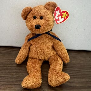 May include: A brown teddy bear with a blue ribbon around its neck. The bear has a Ty Beanie Baby tag attached to its ear. The tag is red with a white star and the words "Beanie Baby" in white.