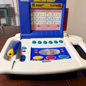 Puede incluir: Un juguete educativo VTech Smart Start Speak & Listen Computer en azul y crema con una pantalla que muestra ecuaciones matemáticas, un lápiz óptico azul y botones coloridos con texto que incluye Plus, Minus, Times, Yes, No, Help, Answer y Repeat.