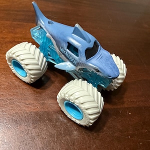 Puede incluir: Un cami&oacute;n monstruo de juguete azul y blanco con un dise&ntilde;o de tibur&oacute;n. El cami&oacute;n tiene neum&aacute;ticos blancos grandes con llantas azules y la palabra "MEGALODON" impresa en el lateral. La carrocer&iacute;a del cami&oacute;n es azul con una aleta de tibur&oacute;n.