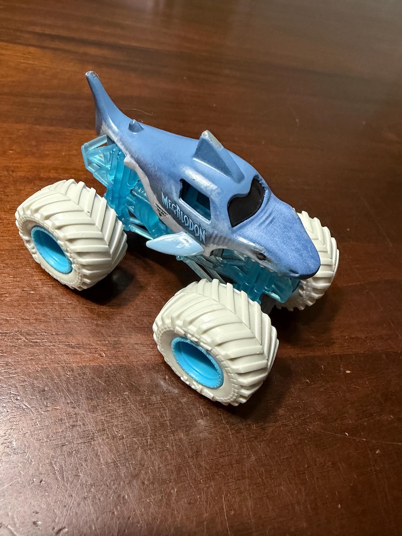 Puede incluir: Un cami&oacute;n monstruo de juguete azul y blanco con un dise&ntilde;o de tibur&oacute;n. El cami&oacute;n tiene neum&aacute;ticos blancos grandes con llantas azules y la palabra "MEGALODON" impresa en el lateral. La carrocer&iacute;a del cami&oacute;n es azul con una aleta de tibur&oacute;n.