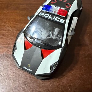 May include: A black, white, and red toy police car with the word "POLICE" on the roof. The toy car has a spoiler and a blue and red light bar. The front of the car has the word "Lamborghini" printed on it.