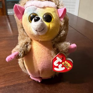 May include: A plush hedgehog toy with large, glittery eyes and a rainbow-colored face. The toy has brown fur, pink ears and paws, and a pink headband. A red heart-shaped tag with the words "Beanie Boos" is attached.