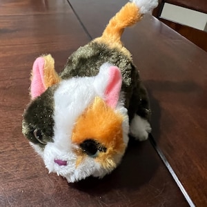 May include: A plush toy cat in a lying position. The cat has a white face with orange and black markings, pink ears, and a pink nose. The body is primarily dark green with orange and white accents. The tail is orange and white.
