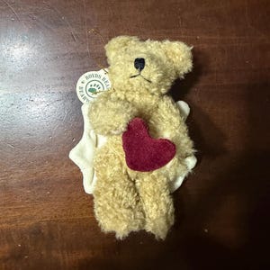 May include: A small, tan-colored Boyd's Bear plush toy with a dark red heart and a white fabric accent. The bear has a black stitched nose and a tag that reads "Boyds Bear." The plush toy is a collectible item.
