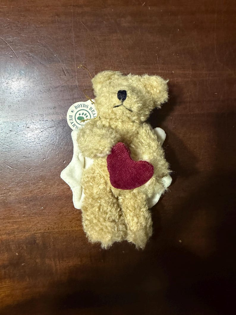 May include: A small, tan-colored Boyd's Bear plush toy with a dark red heart and a white fabric accent. The bear has a black stitched nose and a tag that reads "Boyds Bear." The plush toy is a collectible item.