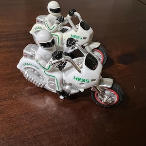May include: Two white toy motorcycles with green and black accents. The motorcycles have the word "HESS" printed on them. The motorcycles are on a brown wooden surface.