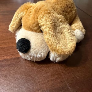 May include: A plush dog toy with tan and cream-colored fur. The dog has a black nose and floppy ears. The toy is lying on a wooden surface.