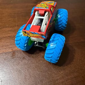 Puede incluir: Un juguete de cami&oacute;n monstruo Hot Wheels con una carrocer&iacute;a estampada en rojo, amarillo y azul y neum&aacute;ticos azules grandes. El cami&oacute;n tiene una jaula antivuelco blanca y el logotipo de Hot Wheels es visible.