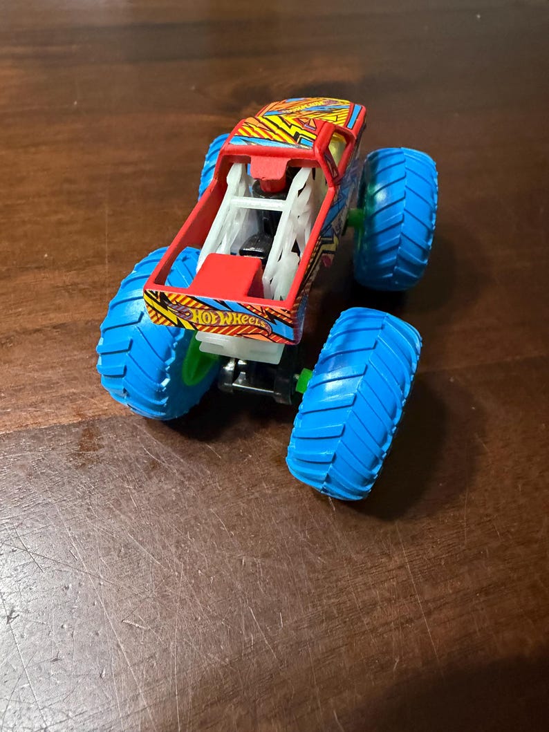 Puede incluir: Un juguete de cami&oacute;n monstruo Hot Wheels con una carrocer&iacute;a estampada en rojo, amarillo y azul y neum&aacute;ticos azules grandes. El cami&oacute;n tiene una jaula antivuelco blanca y el logotipo de Hot Wheels es visible.
