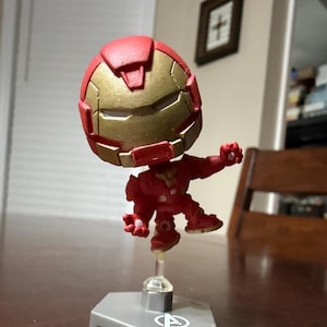 May include: A red and gold Iron Man figurine with a stylized design. The figure is posed mid-action, mounted on a clear stand attached to a gray base. The helmet and chest plate are gold, with the rest of the suit in red.