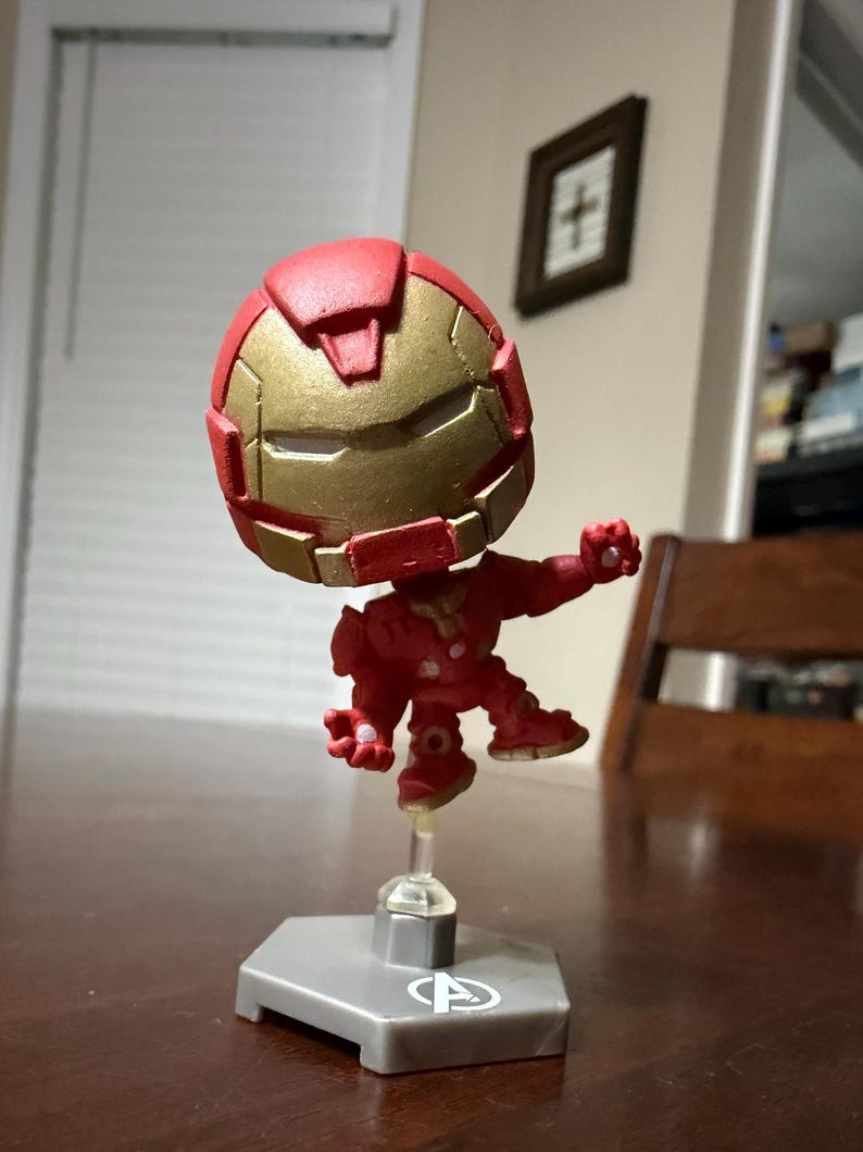 May include: A red and gold Iron Man figurine with a stylized design. The figure is posed mid-action, mounted on a clear stand attached to a gray base. The helmet and chest plate are gold, with the rest of the suit in red.