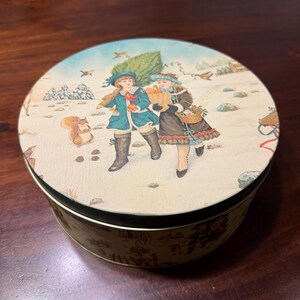 May include: A round metal tin featuring a vintage-style illustration of two children walking through a snowy landscape. One child wears a teal coat and carries a small tree, while the other wears a brown coat and carries a paper bag and basket.