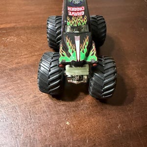 May include: A black toy monster truck with large, black tires. The truck features the words "GRAVE DIGGER" in red and green flames on the hood. The truck has a detailed design with a silver front bumper.