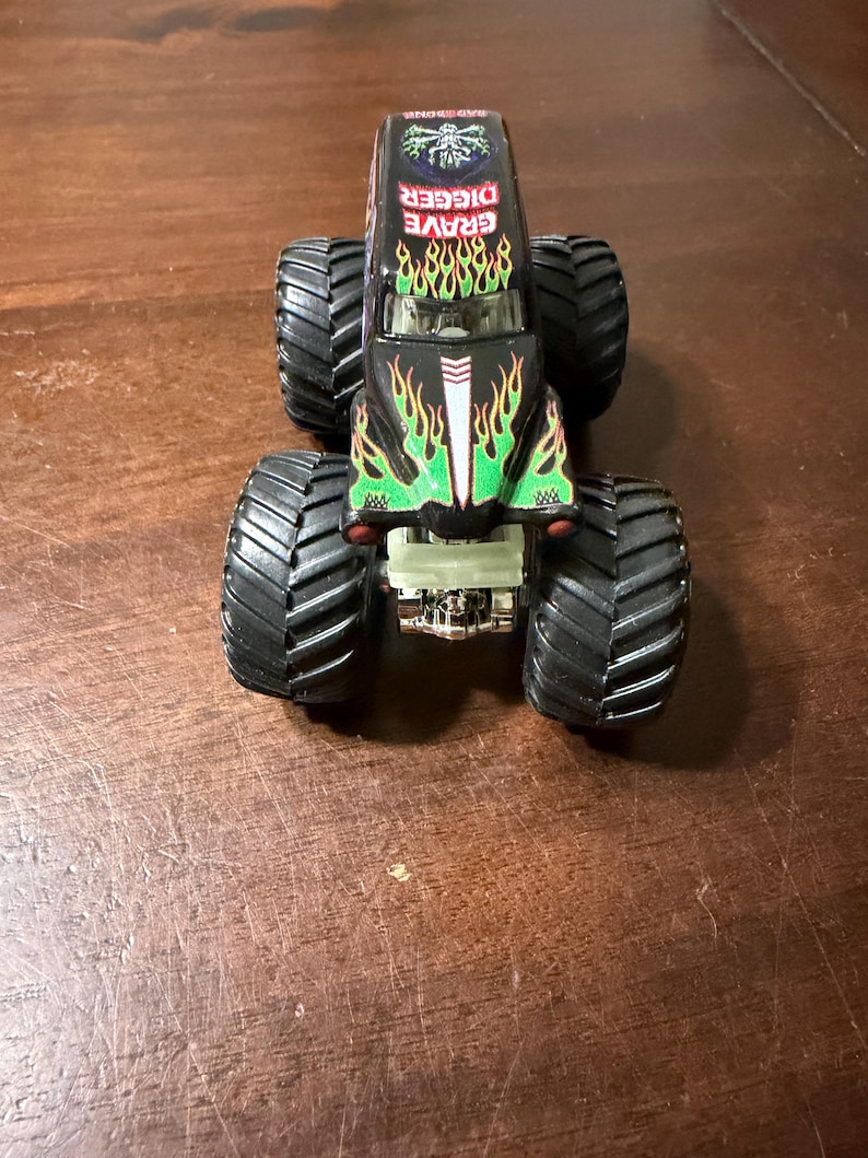 May include: A black toy monster truck with large, black tires. The truck features the words "GRAVE DIGGER" in red and green flames on the hood. The truck has a detailed design with a silver front bumper.