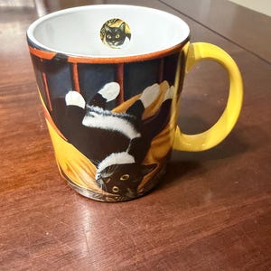 May include: A ceramic mug with a yellow handle and a black and white cat design. The mug features a black cat on a yellow blanket, with a small image of a black cat inside the rim. The mug is on a wooden surface.