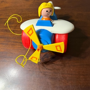 May include: A vintage toy airplane with a yellow propeller and a pull string. The plane has a white body, red wheels, and a blue cockpit. A small figure with yellow hair and an orange shirt sits in the cockpit.