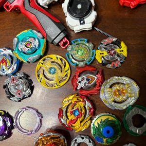 May include: A collection of colorful spinning tops and launchers. The tops feature various designs with red, yellow, blue, green, and silver accents. Launchers are red, white, and black. The items are arranged on a wooden surface.