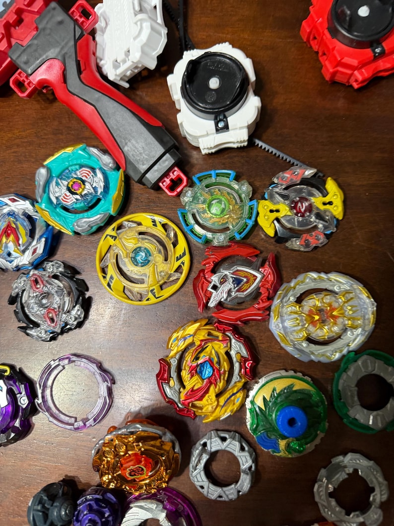 May include: A collection of colorful spinning tops and launchers. The tops feature various designs with red, yellow, blue, green, and silver accents. Launchers are red, white, and black. The items are arranged on a wooden surface.