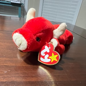 Puede incluir: Un juguete de peluche rojo con forma de toro con cuernos y hocico blancos. El juguete tiene ojos negros y una etiqueta en forma de corazón que dice "Beanie Baby". El juguete está sobre una superficie de madera.