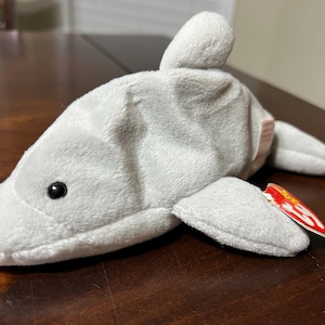 May include: A soft, light grey dolphin toy with a small dorsal fin and flippers. The dolphin has a black eye and a red and yellow tag attached. The toy is made of a plush material.