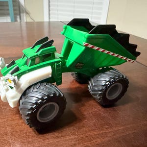 May include: A green and black toy dump truck with large black tires. The truck has white and black accents and a red and white striped detail on the side of the dump bed. The word "DIRT" is visible on the side.