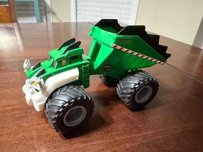May include: A green and black toy dump truck with large black tires. The truck has white and black accents and a red and white striped detail on the side of the dump bed. The word "DIRT" is visible on the side.