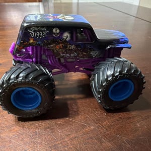 Puede incluir: Un juguete de cami&oacute;n monstruo negro y morado con la inscripci&oacute;n "Grave Digger" en el lateral. El cami&oacute;n tiene neum&aacute;ticos negros grandes con llantas azules y un chasis morado. El cami&oacute;n est&aacute; sobre una superficie de madera.