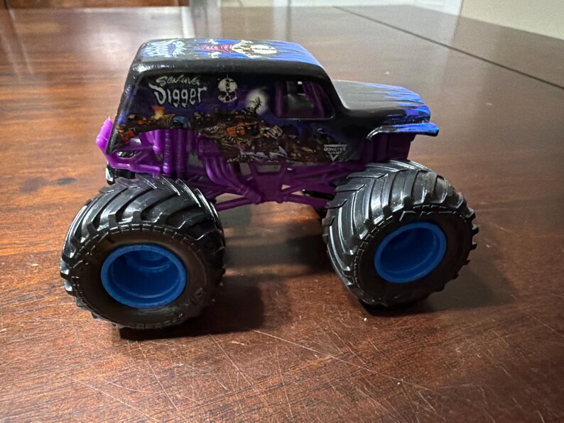 Puede incluir: Un juguete de cami&oacute;n monstruo negro y morado con la inscripci&oacute;n "Grave Digger" en el lateral. El cami&oacute;n tiene neum&aacute;ticos negros grandes con llantas azules y un chasis morado. El cami&oacute;n est&aacute; sobre una superficie de madera.