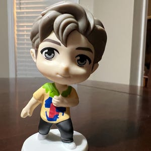 May include: A figurine of a young person with brown hair and large eyes. The figure wears a yellow shirt with a colourful design, black trousers, and white shoes. It stands on a white circular base.
