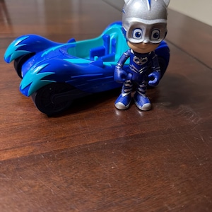 May include: A blue toy car with teal accents and a matching figurine of a character in a blue and silver costume. The toy car has large wheels and a sleek design. The figurine is standing next to the car.