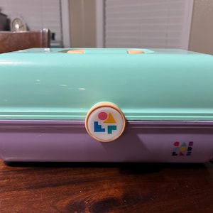 Pode incluir: Uma caixa de armazenamento vintage de duas cores em verde menta e lavanda. A caixa tem um topo arredondado com duas pequenas alças cor de pêssego. Um emblema circular com formas geométricas está na frente. As palavras "CASE MOD" são impressas no canto inferior direito.