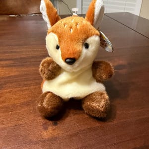 May include: A plush fox toy with orange, brown, and white fur. The fox has large ears, a black nose, and small black eyes. The toy is sitting on a wooden surface.