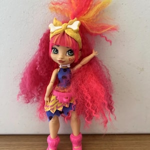 May include: A doll with bright pink, orange, and yellow hair, wearing a yellow headband with a white bow. The doll has a blue top, a skirt with yellow and purple patterns, and pink leg warmers. The doll is standing on a wooden surface.