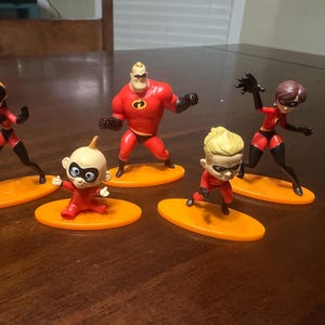May include: Five figurines of the Incredibles family, each standing on an orange oval base. The figures are in dynamic poses, with red and black costumes. The characters include Mr. Incredible, Elastigirl, Violet, Dash, and Jack-Jack.