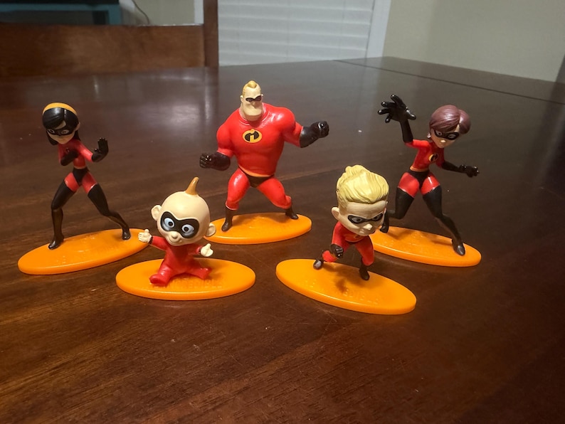 May include: Five figurines of the Incredibles family, each standing on an orange oval base. The figures are in dynamic poses, with red and black costumes. The characters include Mr. Incredible, Elastigirl, Violet, Dash, and Jack-Jack.