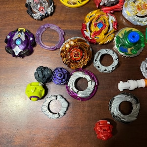 May include: A collection of colorful spinning tops in various designs and colors, including yellow, red, blue, and purple. Some tops have intricate layered designs, while others have simpler patterns. The tops are arranged on a wooden surface.