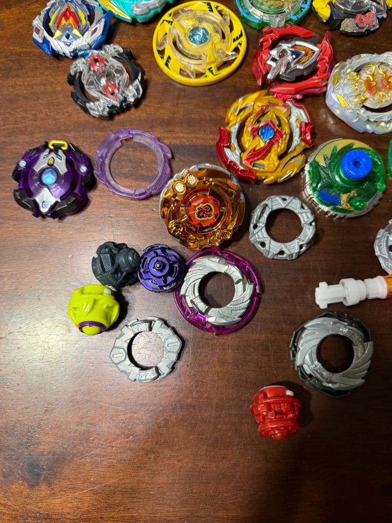 May include: A collection of colorful spinning tops in various designs and colors, including yellow, red, blue, and purple. Some tops have intricate layered designs, while others have simpler patterns. The tops are arranged on a wooden surface.