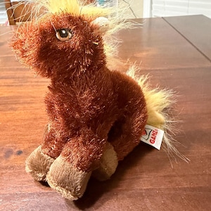 May include: A plush brown horse toy with a shaggy mane and tail. The toy has a light brown muzzle and hooves. A white tag with the brand name "GANZ" is attached to the toy.