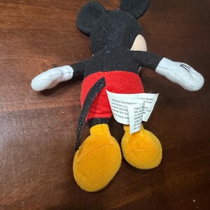 May include: A small Mickey Mouse plush toy. The toy has a black head and ears, a red pair of shorts, yellow feet, and white gloves. A white tag is attached to the toy. The toy is lying on a brown surface.
