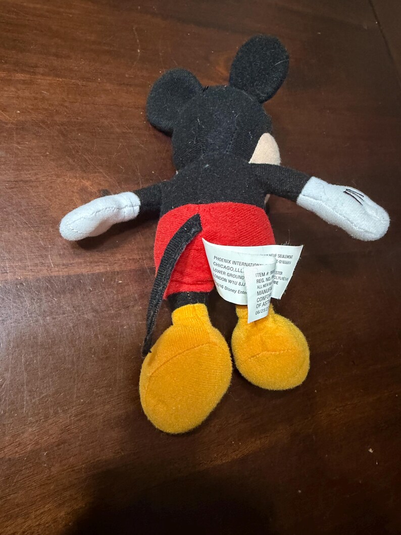 May include: A small Mickey Mouse plush toy. The toy has a black head and ears, a red pair of shorts, yellow feet, and white gloves. A white tag is attached to the toy. The toy is lying on a brown surface.