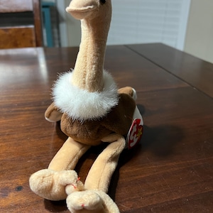 Puede incluir: Un juguete de peluche de avestruz con cuello largo, cuerpo marrón y patas y cabeza color beige. Tiene un collar blanco y esponjoso y una etiqueta roja adjunta. El juguete está sobre una superficie de madera.
