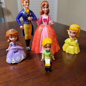 May include: A collection of five plastic figurines of characters from a children's animated series. The figures include a princess in a purple dress, a prince in a blue jacket, and other characters in colorful outfits. The figurines are standing on a wooden surface.