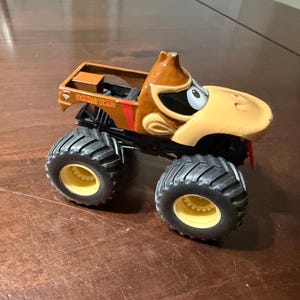 May include: A toy monster truck with a cartoon character's face. It has large black tyres with yellow rims, a brown cab, and the text "DONKEY KONG" on the side.