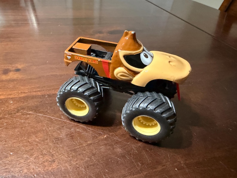 May include: A toy monster truck with a cartoon character's face. It has large black tyres with yellow rims, a brown cab, and the text "DONKEY KONG" on the side.